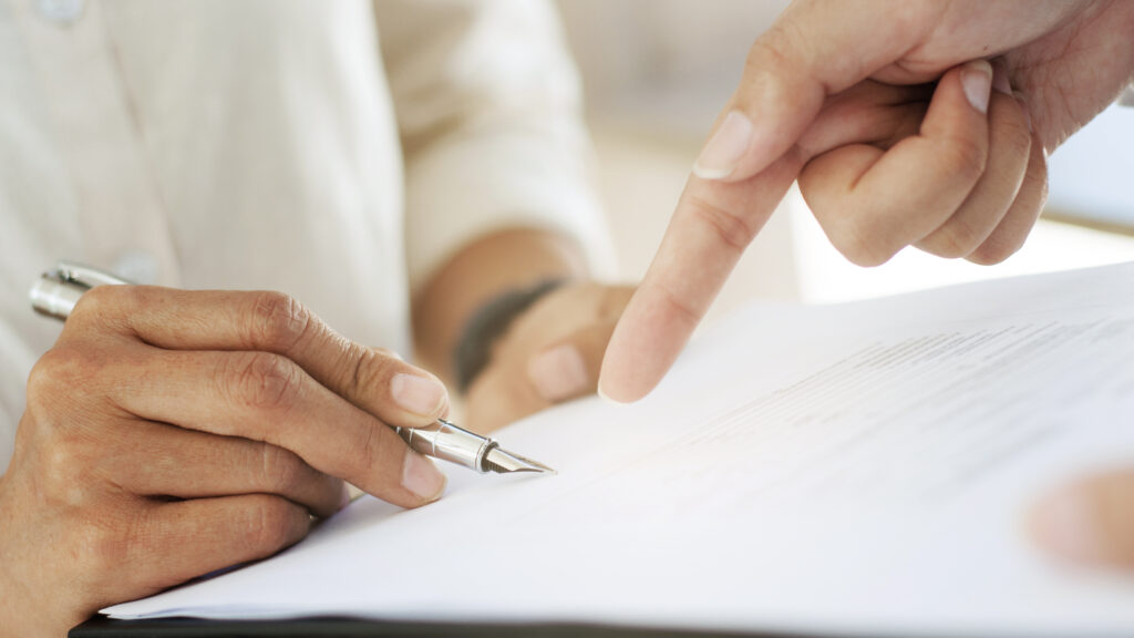 特寫畫面，一人手持鋼筆準備在文件上簽署，另一人用手指指向文件內容進行說明。