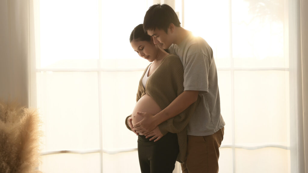 A pregnant woman standing by a bright window, gently holding her bare belly while her partner embraces her from behind.
