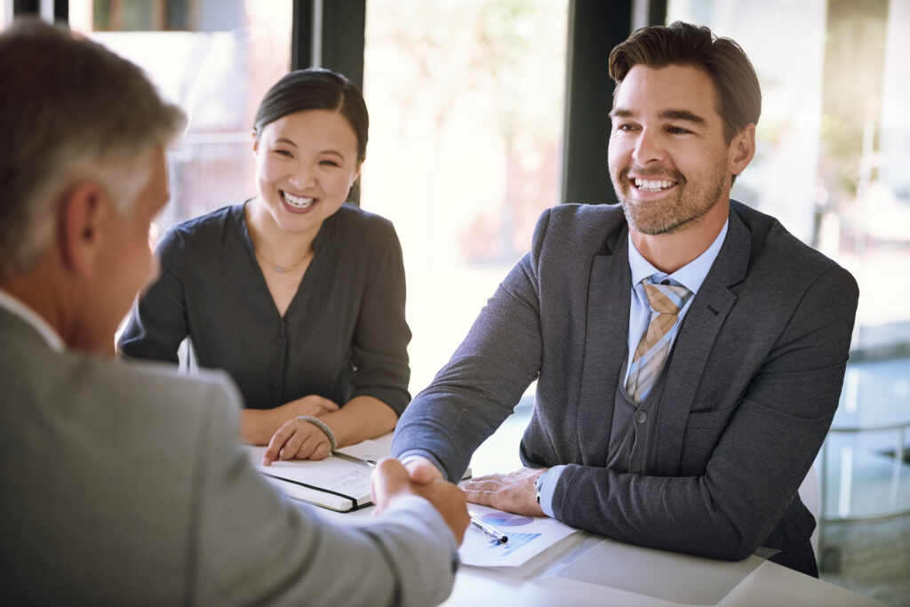 Three adults are gathered for a successful business meeting, sharing smiles and handshakes in a brightly lit urban workplace. Two of the adults are seated at the table while a third adult's arm extends into the frame for a handshake. The woman has dark hair pulled back and wears a dark blouse. One man is wearing a grey suit. Documents on the table suggest financial analysis or strategic planning. The light and airy atmosphere conveys a positive and professional tone. This image could be useful for projects centered on collaboration, finance, career, achievement, and positive business relations. The image's friendly and professional atmosphere could be used for a variety of marketing or corporate communications materials.